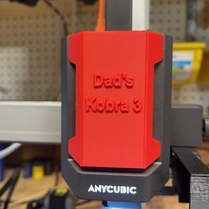 May include: A red and black 3D printer component with the text "Dad's Kobra 3" in raised lettering. The component is attached to a black printer base with the brand name "ANYCUBIC" visible.