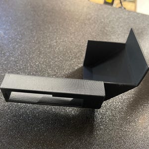 Könnte beinhalten: Ein schwarzes, 3D-gedrucktes Objekt mit einem rechteckigen Rahmen und einer abgewinkelten Rückenstütze. Das Objekt scheint eine Halterung oder Halterung zu sein, möglicherweise für ein Werkzeug oder Gerät. Das Design ist einfach und funktional.