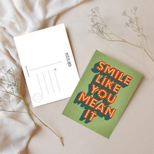 Puede incluir: Dos postales, una en blanco y otra con la frase "SMILE LIKE YOU MEAN IT" en letras naranjas estilo marquesina sobre un fondo verde. La postal en blanco tiene líneas para la dirección y la palabra "POSTCARD" impresa verticalmente.
