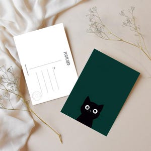 Op de afbeelding: Twee ansichtkaarten, één witte en één donkergroene. De witte ansichtkaart heeft lijnen om te adresseren en het woord "POSTCARD" verticaal afgedrukt. De groene ansichtkaart toont een zwarte kat met grote witte ogen. Gedroogde bloemen staan op de achtergrond.