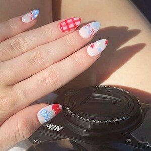 May include: Almond-shaped nails with patriotic designs. The nails feature red, white, and blue patterns, including stars, stripes, and a plaid design. The hand rests near a black camera.