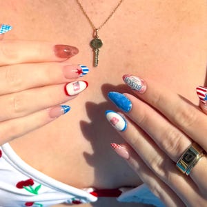 May include: Close-up of a hand with a gold ring and painted nails. The nails feature various designs, including blue and white gingham, red and white stripes, and a Busch beer can. A gold key pendant hangs from a necklace.