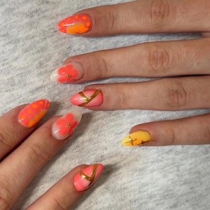 May include: Close-up of a hand with several nails painted with different designs. The nails feature orange and yellow polka dots, floral patterns, and gold accents. The nails are painted in shades of orange, pink, and yellow.