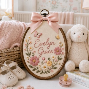 May include: An oval wooden embroidery hoop featuring the name Evelyn Grace stitched in pink thread, surrounded by embroidered flowers and butterflies. A soft pink satin bow is attached to the top of the hoop, which is displayed on a surface.