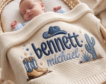 May include: A cream-colored knit baby blanket featuring embroidered designs of a cowboy hat, a pair of cowboy boots, and a cactus. The text "bennett" is embroidered in dark blue, with "michael" below it in a lighter blue script font.