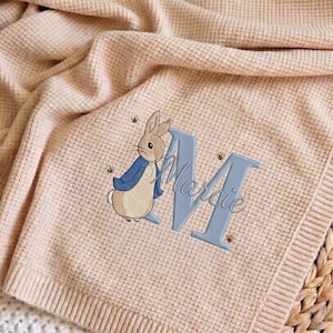 Può includere: Una coperta per neonati in maglia color pesca chiaro con un design di Peter Rabbit. La coperta presenta un personaggio di Peter Rabbit ricamato, una grande lettera blu "M" e il nome "Mardie" in corsivo. La coperta è realizzata in materiale a maglia a nido d'ape.