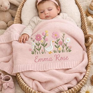 May include: A soft pink knitted baby blanket featuring an embroidered white goose surrounded by pink and yellow wildflowers, with the name Emma Rose stitched in cursive script. The blanket is draped over a sleeping infant in a woven wicker basket.