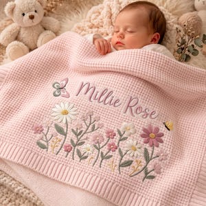 Puede incluir: Una manta de bebé de punto gofrado rosa claro con diseños florales bordados en blanco, rosa y verde, una pequeña mariposa y el nombre Millie Rose cosido en letra cursiva. Un osito de peluche y un conejo de juguete son visibles cerca.