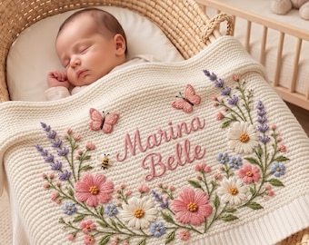 May include: A cream-colored knit baby blanket featuring embroidered floral motifs, including pink and white flowers, lavender sprigs, two butterflies, and a small bee. The name Marina Belle is embroidered in pink script in the center.