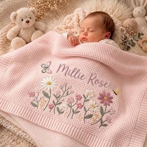 May include: A light pink waffle-knit baby blanket featuring embroidered floral designs in white, pink, and green, with a small butterfly and the name Millie Rose stitched in cursive script. A plush teddy bear and a rabbit toy are visible nearby.