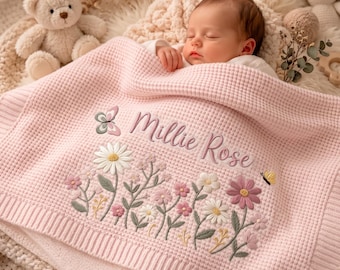 May include: A light pink waffle-knit baby blanket featuring embroidered floral designs in white, pink, and green, with a small butterfly and the name Millie Rose stitched in cursive script. A plush teddy bear and a rabbit toy are visible nearby.