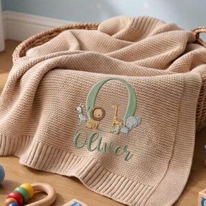 Könnte beinhalten: Eine beige gestrickte Babydecke mit geripptem Rand, verziert mit einem gestickten grünen Buchstaben O, einem Zebra, einem Löwen, einer Giraffe und einem Elefanten, wobei der Name Oliver darunter in grünem Garn gestickt ist, in einem Weidenkorb.