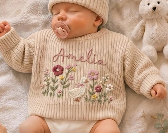 May include: A cream-colored ribbed knit sweater featuring the embroidered name Amelia above a floral and duck design, paired with a matching beanie and cuffed socks. A plush white rabbit toy and a small wooden toy car are placed nearby.