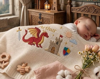 May include: A cream-colored knit baby blanket featuring embroidered designs of a red dragon, a stone castle, a sword, a shield, a sun, and a cloud, with the name Arthur stitched in gold thread. The blanket is decorated with wooden toys.