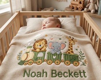 May include: A cream-colored knit baby blanket featuring an embroidered green train carrying a lion, an elephant, and a giraffe. The blanket is personalized with the name Noah Beckett in green text and includes small cloud and star accents.