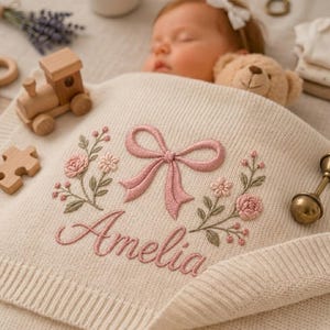 May include: A cream-coloured knit baby blanket featuring an embroidered design of a pink bow, floral sprigs, and the name Amelia in pink script. The blanket is displayed with a wooden toy train, a puzzle piece, and a brass rattle nearby.
