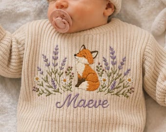 May include: A cream-colored ribbed knit sweater featuring an embroidered design of a fox surrounded by lavender sprigs and small white flowers. The name Maeve is embroidered in purple cursive script below the fox. The sweater has a matching beanie.