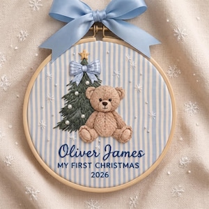May include: A round wooden embroidery hoop featuring light blue and white striped fabric. It displays a stitched brown teddy bear, a green Christmas tree with a star, and the text Oliver James MY FIRST CHRISTMAS 2026. A light blue ribbon bow is attached at the top.