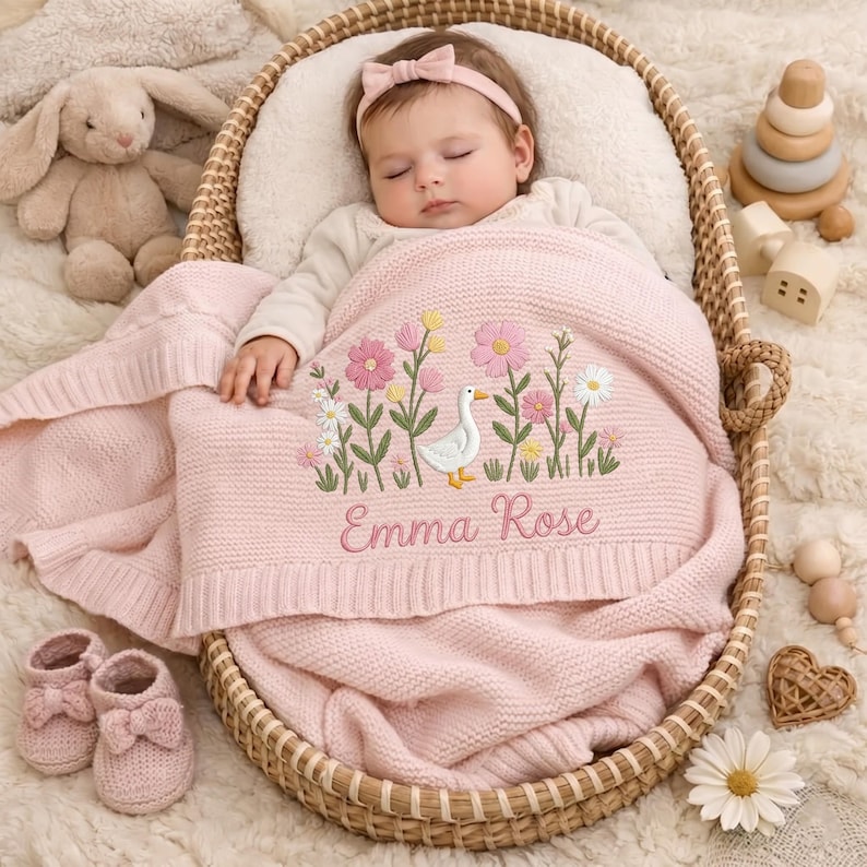 May include: A soft pink knitted baby blanket featuring an embroidered white goose surrounded by pink and yellow wildflowers, with the name Emma Rose stitched in cursive script. The blanket is draped over a sleeping infant in a woven wicker basket.