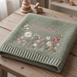 May include: A sage green knitted blanket featuring a waffle texture and a ribbed border. The center is embroidered with the name Millie Rose in pink script, surrounded by a colorful floral arrangement of daisies and wildflowers with a small butterfly.