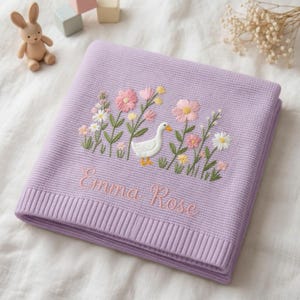 May include: A lavender knitted baby blanket featuring an embroidered scene of a white goose surrounded by pink and yellow flowers. The name Emma Rose is embroidered in pink cursive script below the floral design. The blanket has a ribbed border.