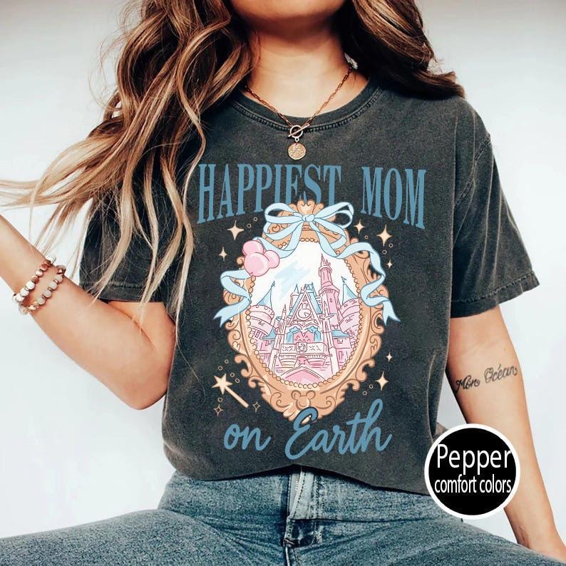 Puede incluir: Camiseta gris oscuro con el texto "Happiest Mom on Earth" en azul. La camiseta presenta un gr&aacute;fico de un castillo dentro de un marco adornado con un lazo y estrellas. La camiseta es de Pepper comfort colors.