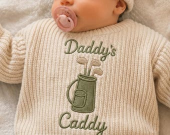 May include: A cream-colored ribbed knit sweater featuring an embroidered design of a golf bag with clubs and the text Daddy's Caddy in dark green thread. The sweater is worn by an infant wearing a matching cream-colored knit beanie and a pink pacifier.