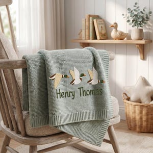 May include: A sage green knitted baby blanket featuring an embroidered design of three flying ducks in shades of white, brown, and green. The name Henry Thomas is embroidered in dark green thread below the ducks on the textured fabric.