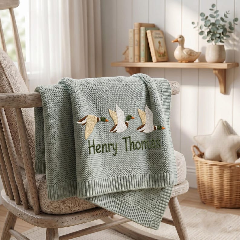 May include: A sage green knitted baby blanket featuring an embroidered design of three flying ducks in shades of white, brown, and green. The name Henry Thomas is embroidered in dark green thread below the ducks on the textured fabric.