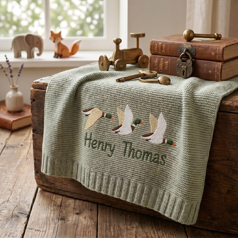May include: A sage green knitted baby blanket featuring an embroidered design of three flying ducks and the name Henry Thomas stitched in dark green thread. The blanket has a ribbed border and is draped over a rustic wooden chest.