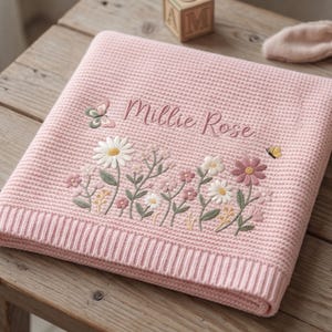 May include: A folded light pink waffle-knit blanket featuring embroidered floral designs in white, pink, and green, along with a small butterfly. The name Millie Rose is embroidered in a cursive script above the flowers on the textured fabric.
