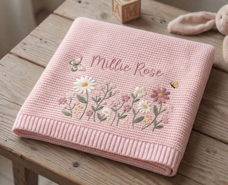 May include: A folded light pink waffle-knit blanket featuring embroidered floral designs in white, pink, and green, along with a small butterfly. The name Millie Rose is embroidered in a cursive script above the flowers on the textured fabric.