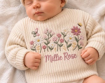 May include: A cream-colored ribbed knit sweater and matching beanie set featuring an embroidered floral design with pink, white, and green flowers, butterflies, and the name Millie Rose stitched in pink script across the chest area.