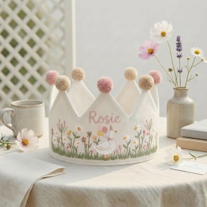 Puede incluir: Una corona de tela color crema con un diseño bordado floral y de ganso. La corona tiene pompones rosas y beige y el nombre "Rosie" bordado en rosa. La corona está sobre un mantel blanco.
