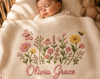 May include: A cream-colored knit baby blanket featuring an embroidered design of various wildflowers, a butterfly, and a bee. The name Olivia Grace is embroidered in pink cursive script at the bottom center of the blanket, which is draped over a sleeping infant.