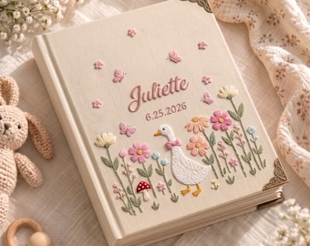 May include: A cream-colored fabric-covered photo album featuring embroidered details of a white goose, pink and yellow flowers, butterflies, and a red mushroom. The name Juliette and the date 6.25.2026 are embroidered in pink. It has decorative metal corners.