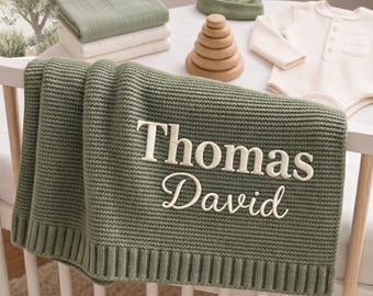 May include: A sage green knitted baby blanket featuring the embroidered name Thomas David in white lettering. The blanket is draped over a white crib, accompanied by a stack of folded blankets, a matching beanie, a wooden stacking toy, and a cream bodysuit.