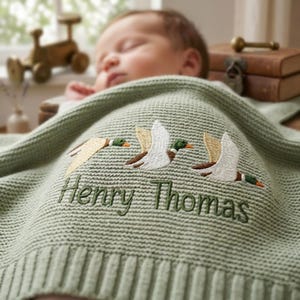 May include: A sage green knitted baby blanket featuring an embroidered design of three flying mallard ducks in white, brown, and green thread. Below the ducks, the name Henry Thomas is embroidered in dark green script on the textured fabric.