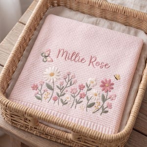 May include: A light pink waffle-knit blanket featuring the embroidered name Millie Rose in a cursive script. The blanket is decorated with a colorful floral garden motif including daisies, small pink flowers, a butterfly, and a tiny bee.