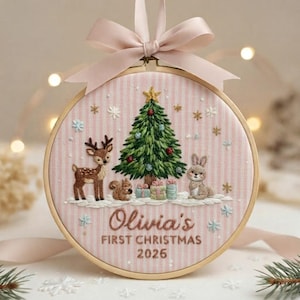 May include: A round wooden embroidery hoop featuring a pink and white striped fabric background. The design includes a green Christmas tree, a deer, a squirrel, a rabbit, and gifts. Text reads: Olivia's FIRST CHRISTMAS 2026. A pink ribbon bow is attached to the top.