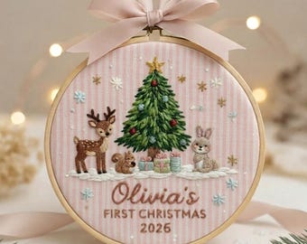 May include: A round wooden embroidery hoop featuring a pink and white striped fabric background. The design includes a green Christmas tree, a deer, a squirrel, a rabbit, and gifts. Text reads: Olivia's FIRST CHRISTMAS 2026. A pink ribbon bow is attached to the top.