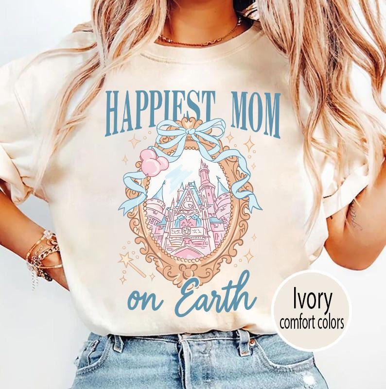 Puede incluir: Camiseta color marfil con la frase "Happiest Mom on Earth" en azul. El dise&ntilde;o incluye un marco decorativo con una ilustraci&oacute;n de castillo rosa y azul, y un lazo. El texto "Ivory comfort colors" est&aacute; en un c&iacute;rculo.