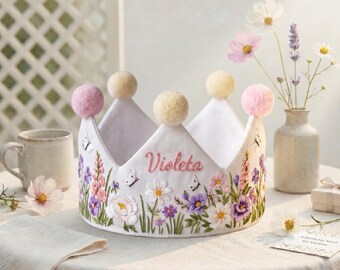 Könnte beinhalten: Eine weiße Stoffkrone, bestickt mit bunten Blumen, Schmetterlingen und dem Namen "Violeta" in Rosa. Die Krone hat drei gelbe und rosa Pompons oben. Eine Geburtstagskarte und Blumen in einer Vase sind im Hintergrund.