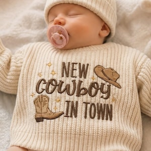 May include: A cream-colored ribbed knit sweater featuring brown embroidered text that reads NEW COWBOY IN TOWN, accompanied by an embroidered brown cowboy hat and a single cowboy boot, accented with small gold star shapes.