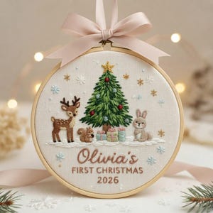 May include: A circular embroidery hoop ornament featuring a cream fabric background with a hand-stitched Christmas tree, a deer, a squirrel, and a rabbit. The text reads Olivia's FIRST CHRISTMAS 2026. A light pink satin ribbon is tied at the top.