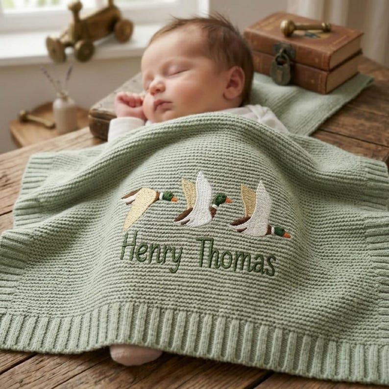 May include: A sage green knitted baby blanket featuring an embroidered design of three flying ducks in white, brown, and green thread. The name Henry Thomas is embroidered in dark green script below the ducks. The blanket has a ribbed border.