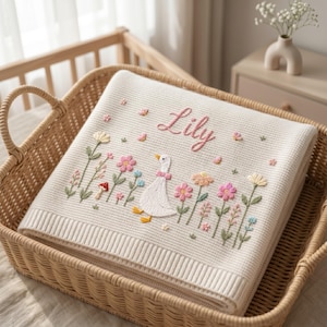 May include: A cream-coloured knit blanket featuring embroidered floral motifs, a small mushroom, and a white goose wearing a pink bow. The name Lily is embroidered in pink script at the top. The blanket has a ribbed border and a textured waffle-knit pattern.
