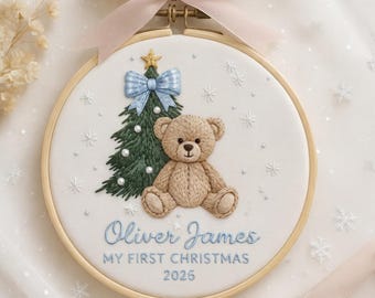 May include: A circular embroidery hoop featuring a light brown teddy bear sitting next to a green Christmas tree with a blue bow and gold star. The white fabric is embroidered with the text Oliver James, My First Christmas, 2026, and snowflakes.