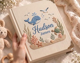 May include: A cream-colored photo album featuring embroidered ocean-themed designs including a blue whale, a green turtle, coral, seashells, and seagulls. The name Hudson James is embroidered in blue script in the center with gold metal corners.
