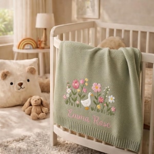 May include: A sage green knitted baby blanket featuring an embroidered scene of a white duck surrounded by pink and yellow wildflowers. The name Emma Rose is embroidered in pink cursive script below the floral design on the textured fabric.
